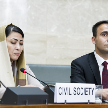 A woman with a head scarf and a man are sitting in a conference room with serious faces. The signs in front of them read "civil society". 