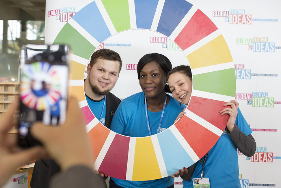 Three young persons holding up a life-size SDG wheel and looking into a mobile phone camera through the middle of the wheel.