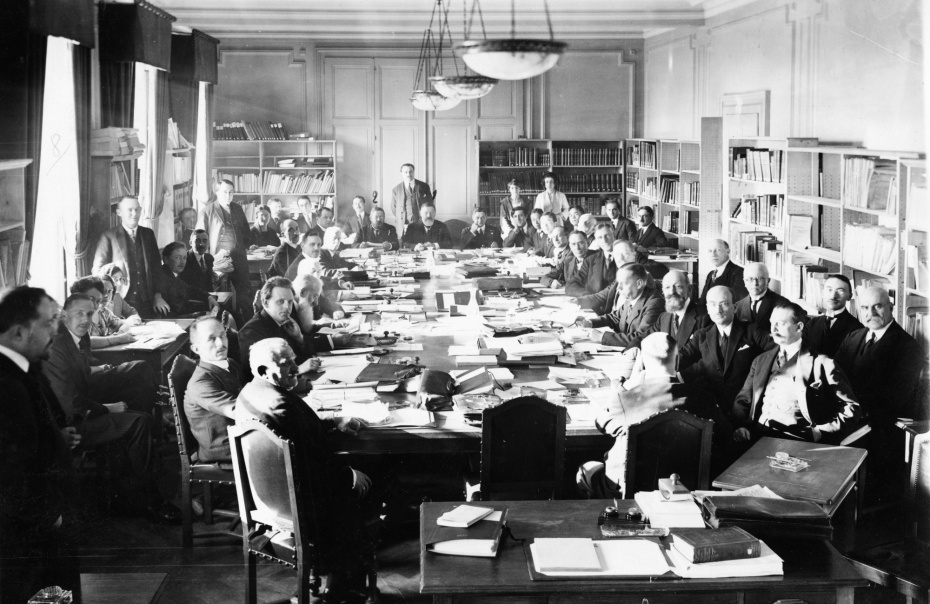 A black and white image of a group of men working around a large table in a room full of book shelves. 