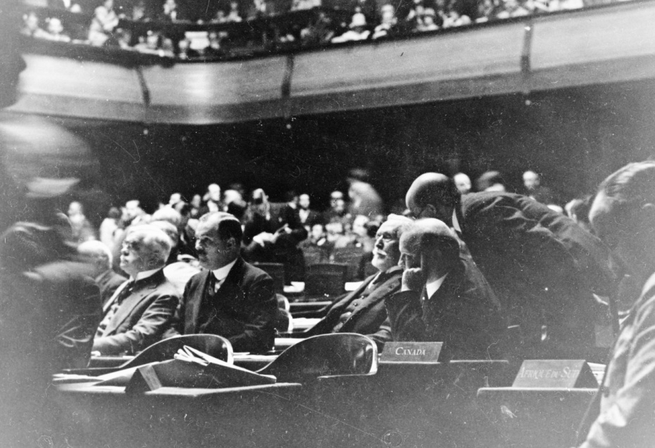 A group of men sitting inside a conference hall, listening. The photo is in black and white. 