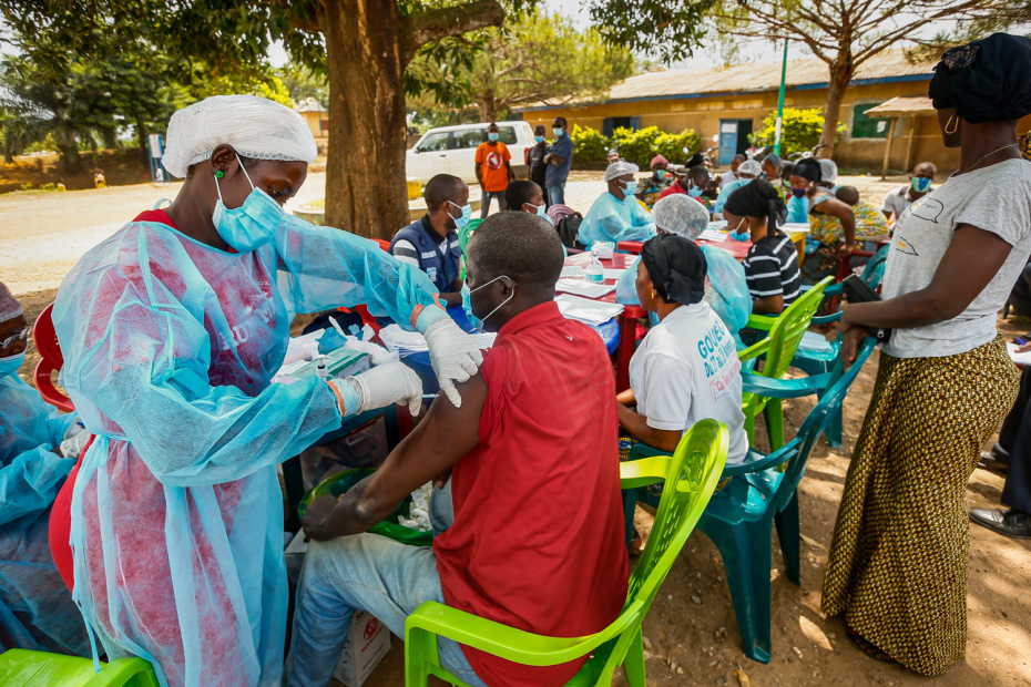 A health worker injects a patient in an outdoor setting. A group of people is sitting at a long table behind the two.
