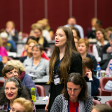 A young woman standing in the middle of a crowded conference room, speaking.