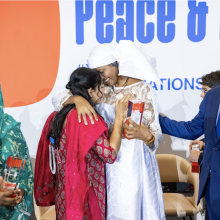 Two women in the center of the image give each other a hug. On a banner at the back, one can read the word "peace".