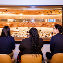 Four visitors are seen from the back as they sit in the gallery of meeting room XIX, following the ongoing meeting.