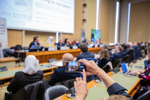 A person taking a photo on a mobile phone of a meeting taking place. Only the phone screen is clear, the rest of the image is blurry. 