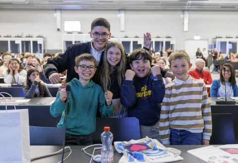 4 children and a young man, laughing and smiling at the camera. Behind them, a meeting room full of young people.