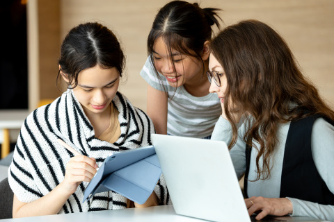 Three young women leaning over their computers, engaged in a task.