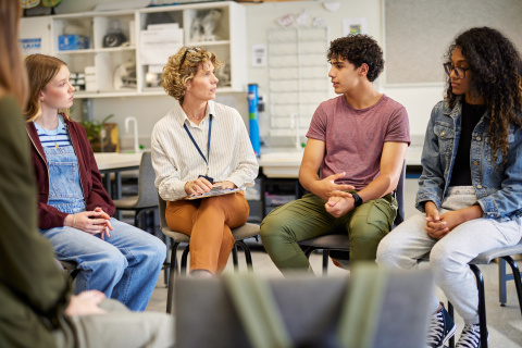 A group of young people sitting in a circle with a middle-aged woman, discussing.