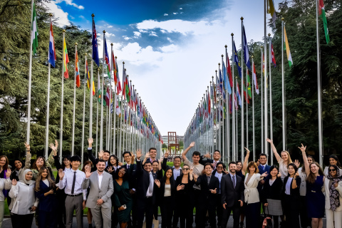 A group of young people standing at the alley of flags outside the Palais des Nations. In the background, one can see the iconic broken chair statue.