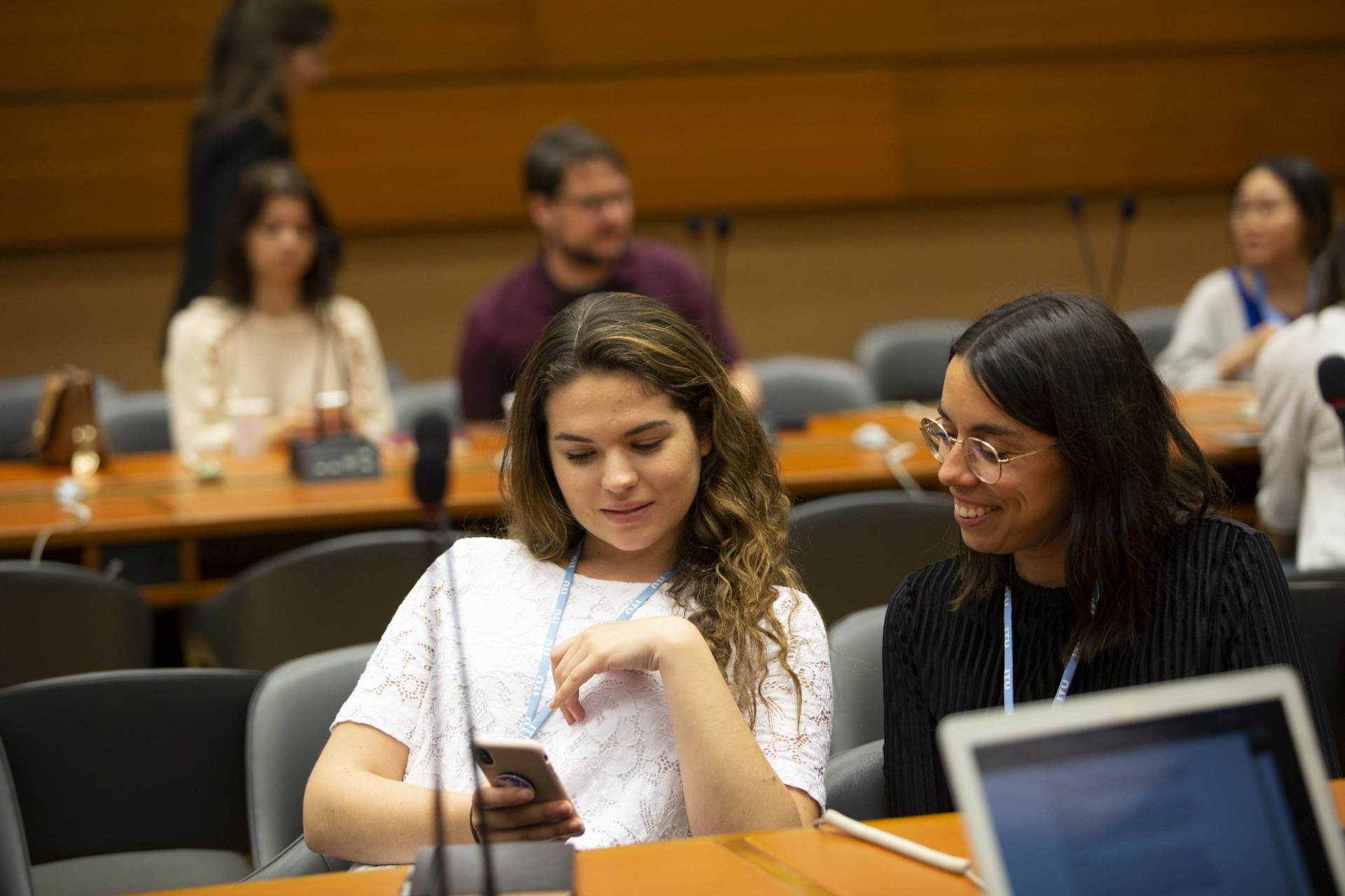 Two young women sitting inside a UN conference room, smiling at something they see on a mobile phone