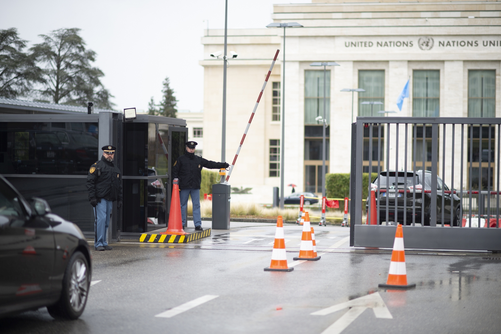 Two UN Security staff are standing at one of the entrance gates at the Palais des Nations. A car is about to enter, the gate is open. The background shows one of the UN buildlings