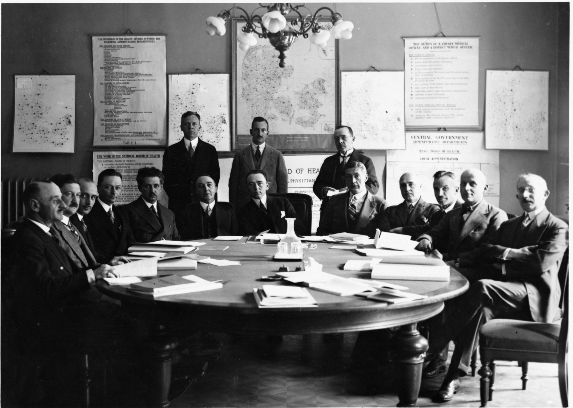 A group of men sitting around a table in a black and white photograph. On the wall behind the group are several large posters and maps. 