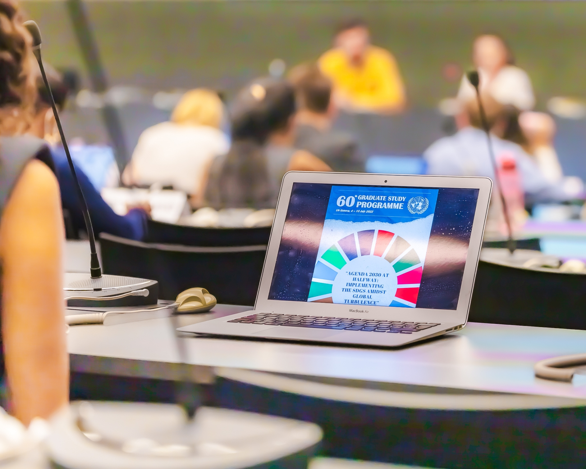 A laptop shows the Graduate Study Programme banner. In the background, students sit in a meeting room and follow a discussion. The persons are blurred. 