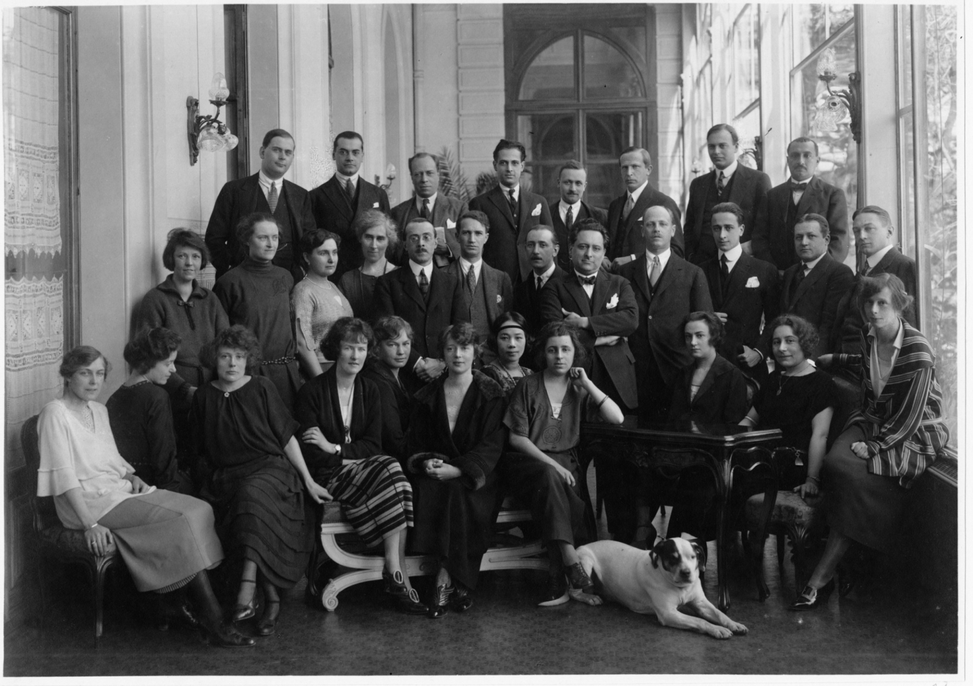 A group of women and men posing for a black and white group picture in a large room with high ceilings. In the first row, there is a dog as well. 