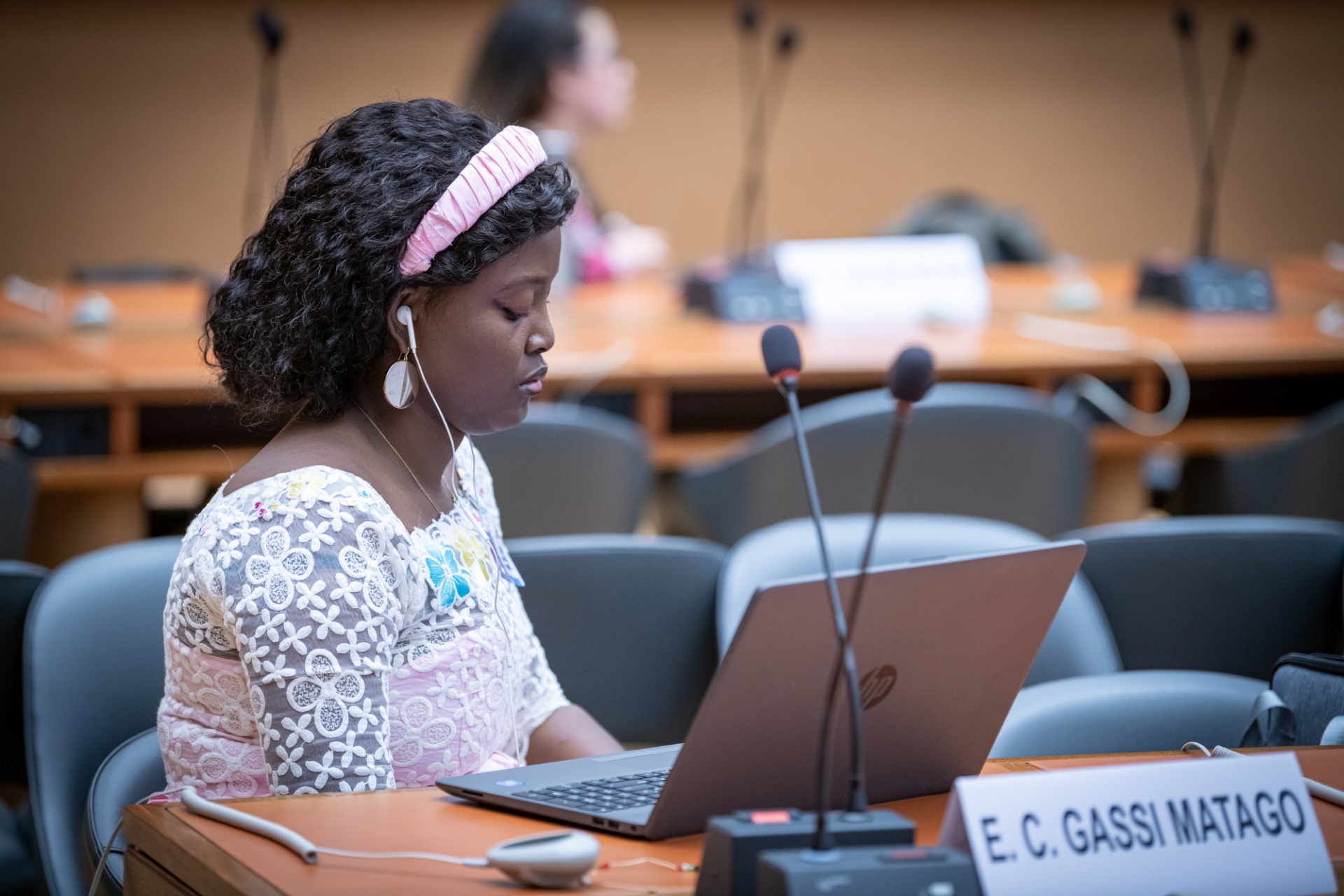 A woman sitting with her laptop inside a meeting room. She has earphones on and listens attentively.