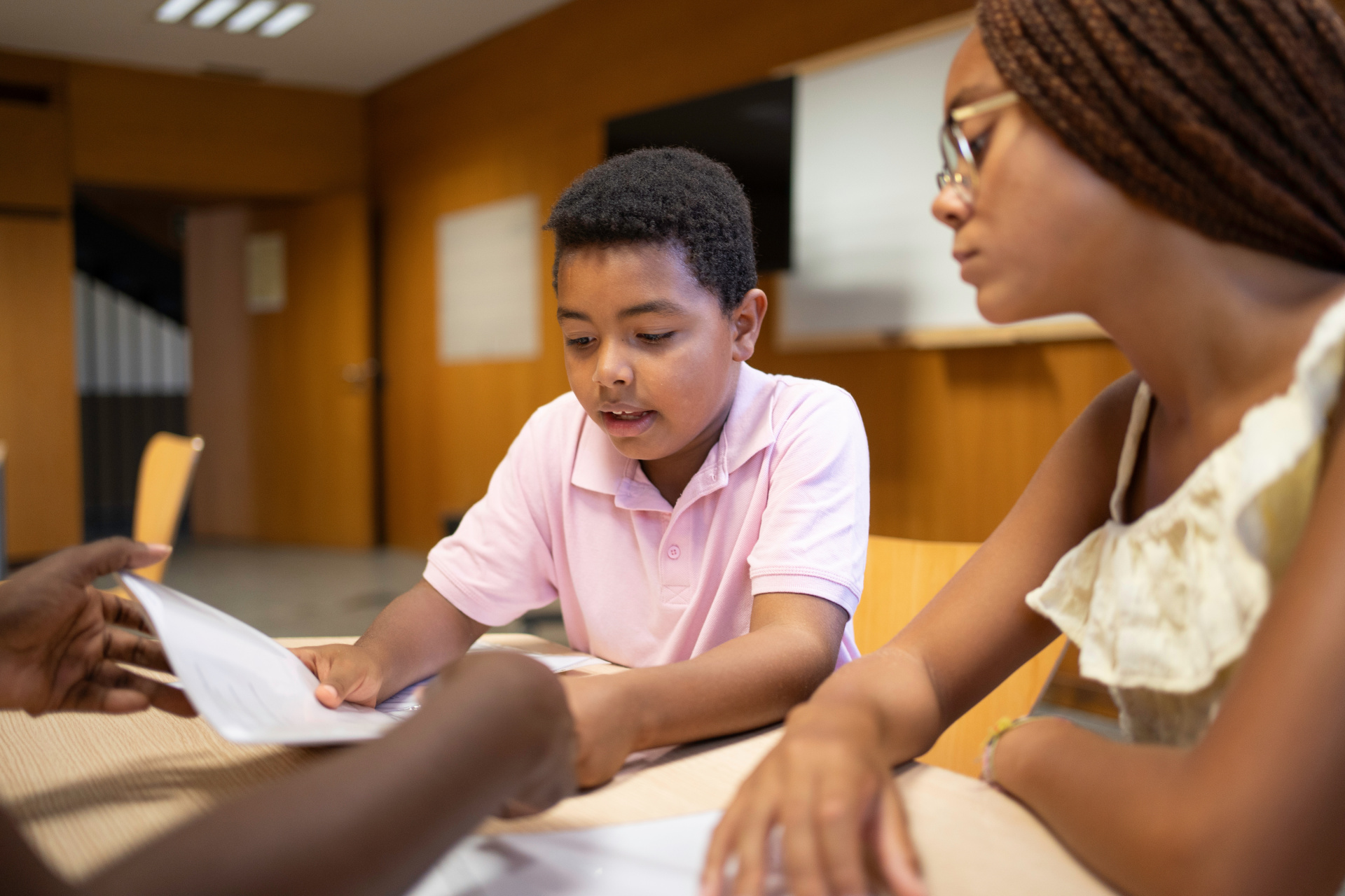 A boy and a girl sitting at a table, discussing a paper. Hands of a third person reach inside the picture from the off.