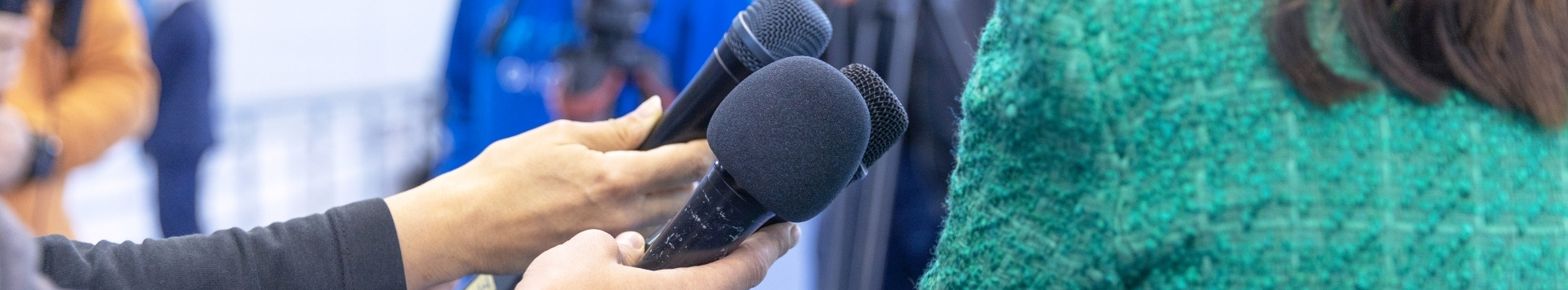 A woman, shown from the back is answering questions to the media. A number of cameras and microphones point in her direction. 