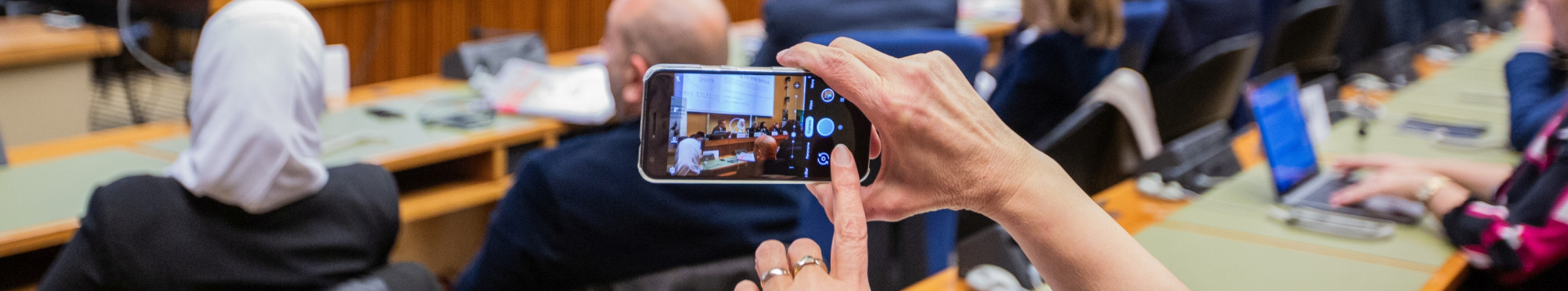 A person taking a photo on a mobile phone of a meeting taking place. Only the phone screen is clear, the rest of the image is blurry.