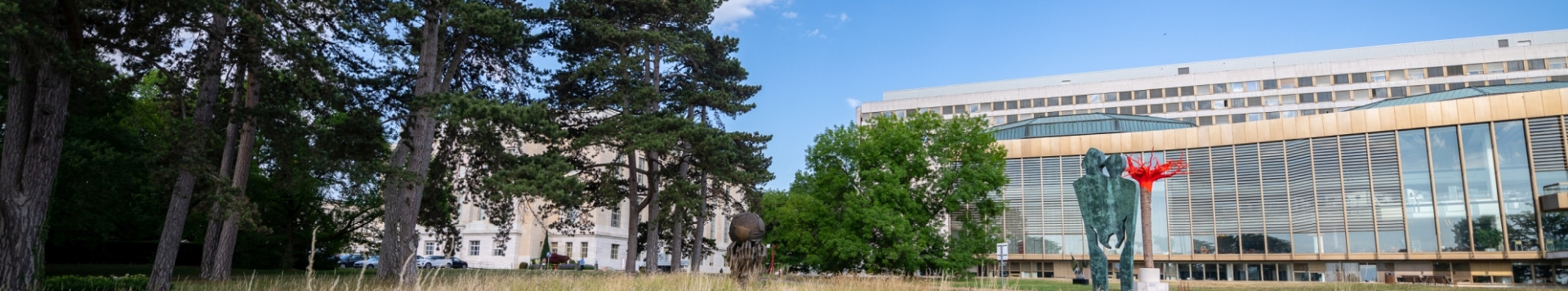 A panoramic view of one of the buildings inside the Palais des Nations next to some trees and behind some artwork. 