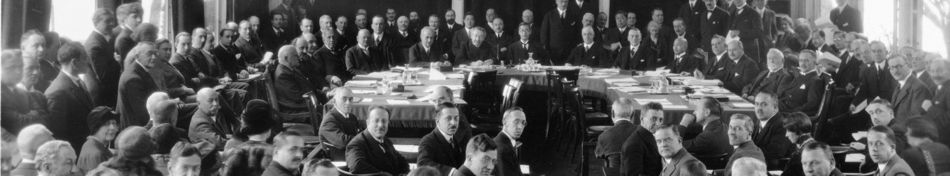 A black and white photography of the League of Nations Council: Three rows of delegate are gathered around a large table, all looking towards the camera. 