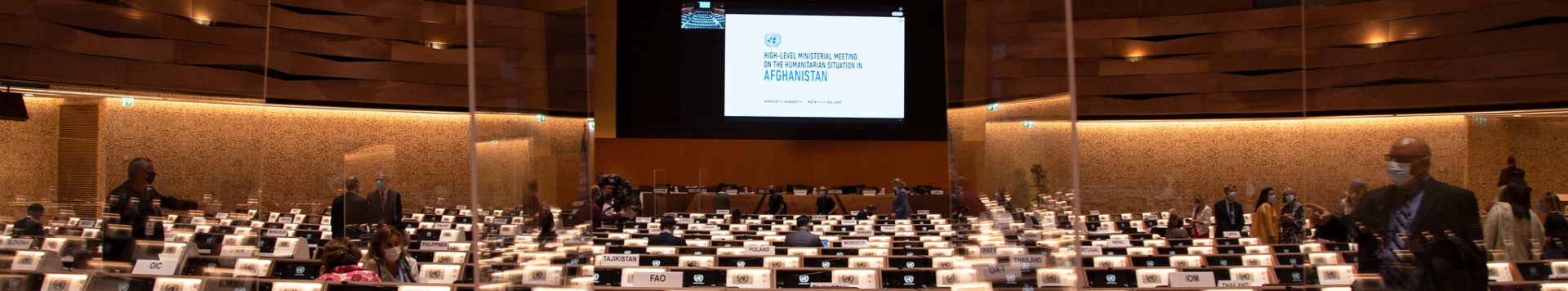 An almost empty conference room, some people are searching seats. A big screen in front shows the title of the event,