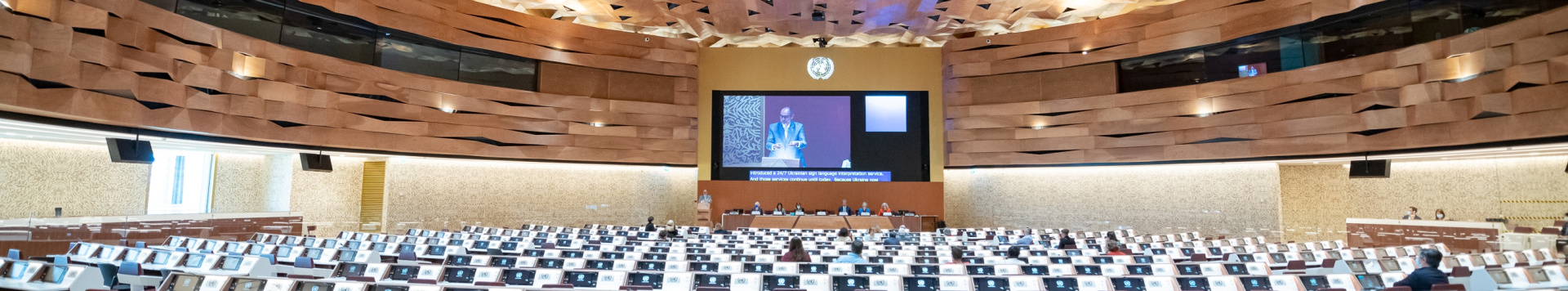 A very large screen projects a speaker behind the panel members in one of the UN conference rooms. 