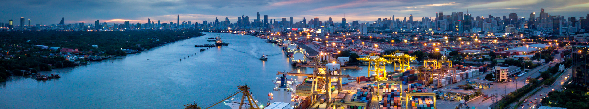 A port scene with cranes, ships and containers next to a river at night. In the background, the skyline of a larger city is seen.