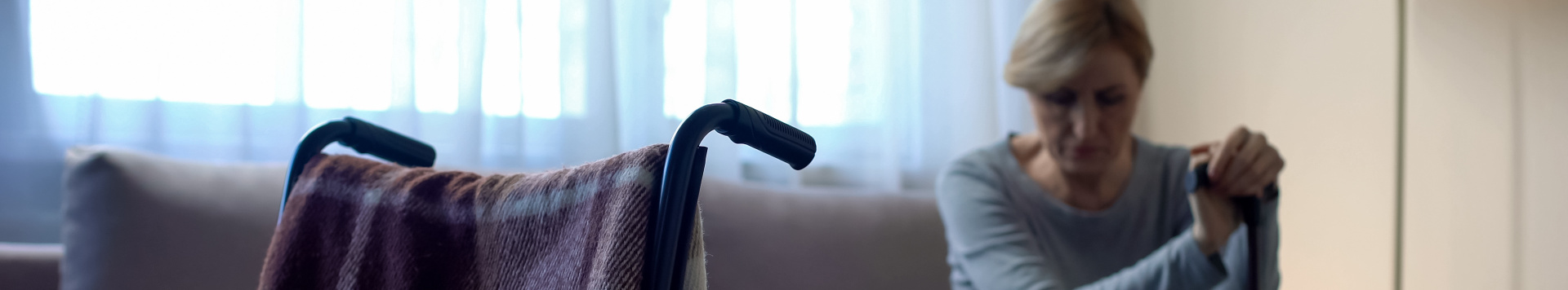 A wheelchair on the left in the foreground, and a woman, our of focus, with a stick sitting on a sofa in the background, looking down.